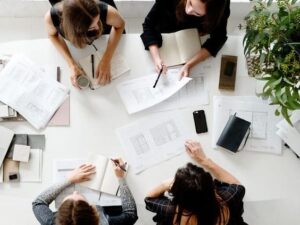 Empat orang sedang berdiskusi di sekitar meja kerja berwarna putih yang dipenuhi gambar rancangan, buku catatan, alat tulis, ponsel, dan tanaman hias.